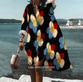 A woman in a black and red floral dress stands by the water, gazing thoughtfully at the serene landscape.